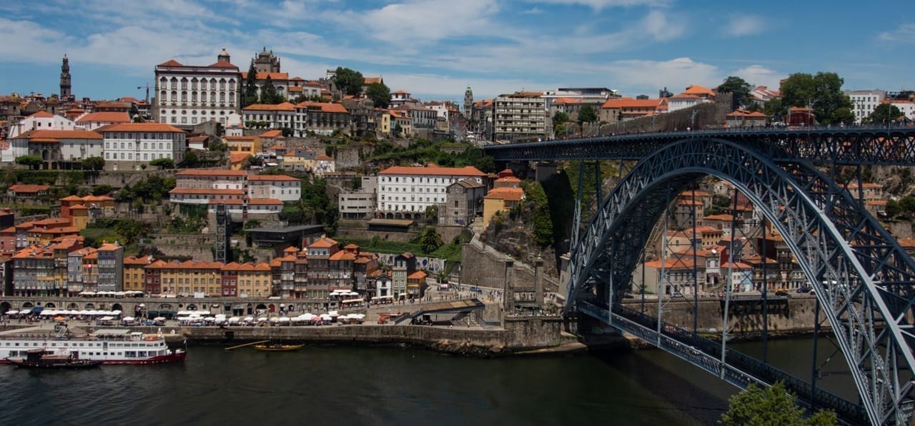 Porto's Ribeira waterfront at blue hour, six-story azulejo-tiled buildings glowing under the Dom Luis I bridge, port wine cellars across the Douro in Vila Nova de Gaia